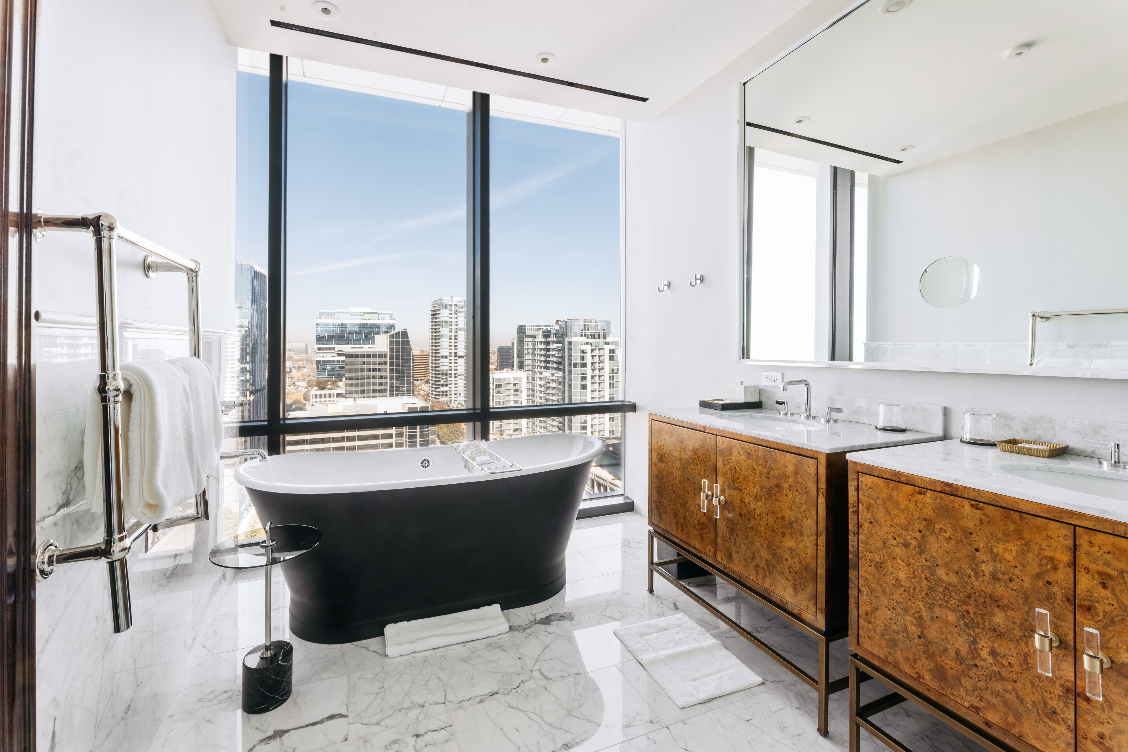 A modern bathroom with a freestanding bathtub, a large mirror, and a view of the city through floor-to-ceiling windows.