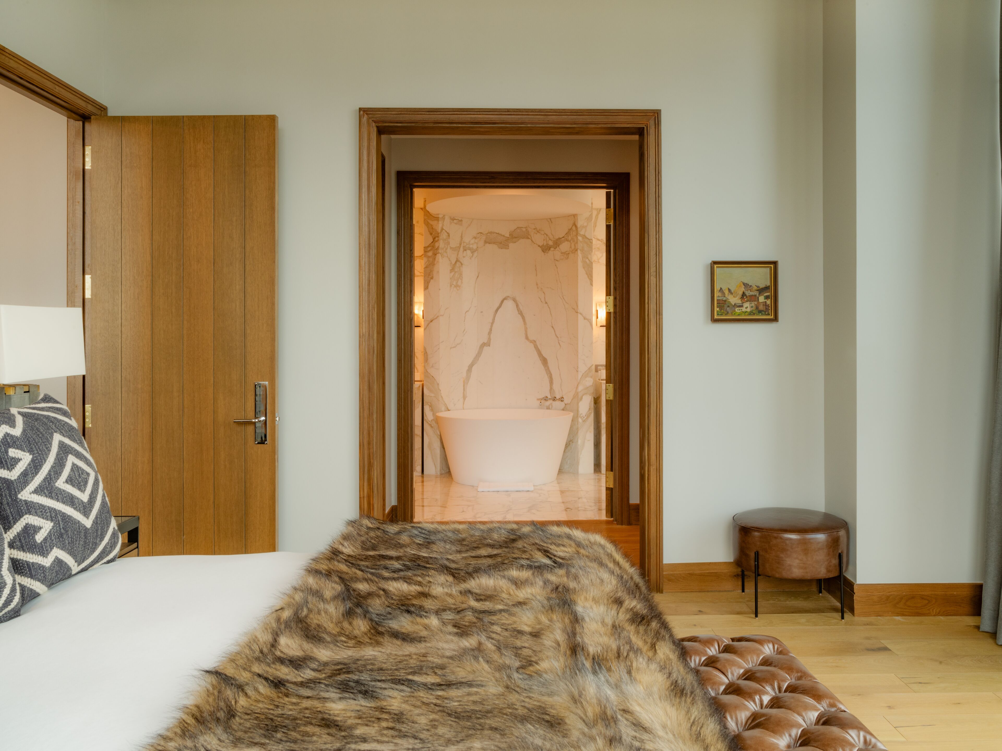 A bedroom with a large bed, a wooden door, a bathroom with a bathtub, and a small stool.