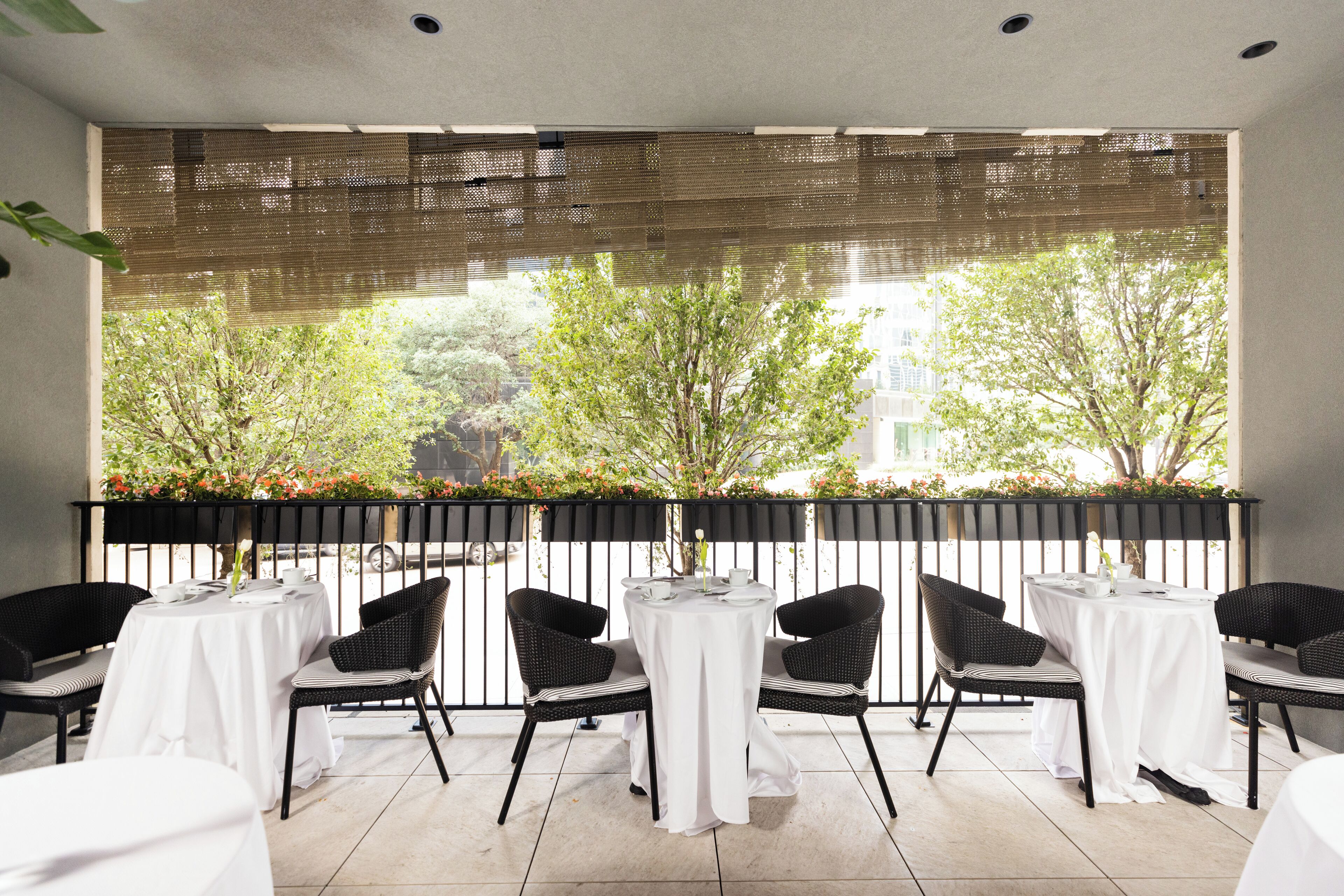 A terrace with tables and chairs, a view of trees and buildings, and a black metal railing.