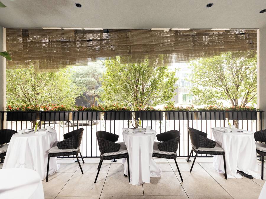 A terrace with tables and chairs, a view of trees and buildings, and a black metal railing.