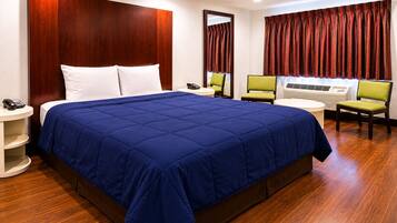 A hotel room with a large bed, a bedside table with a phone, a chair, a desk with a computer, and a window with curtains.