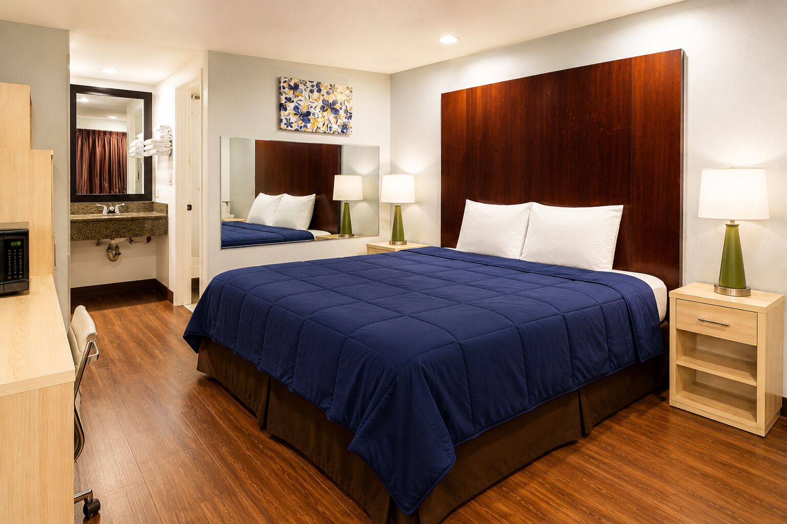 A hotel room with a large bed, wooden headboard, bedside tables with lamps, a mirror, a bathroom with a shower, and a wooden floor.