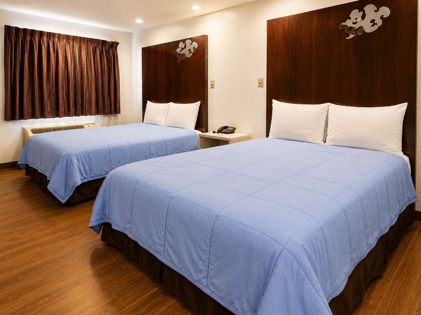 A hotel room with two beds, wooden flooring, a curtain, and a wall-mounted phone.