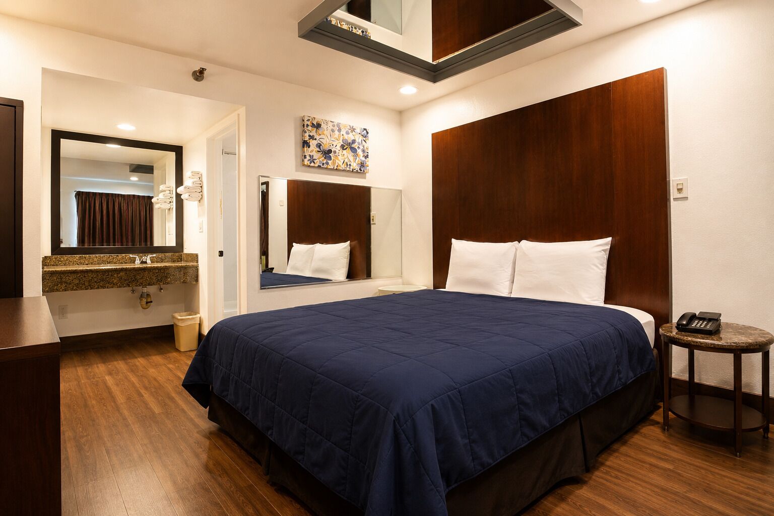 A hotel room with a large bed, a wooden headboard, a bedside table with a phone, a mirror, and a bathroom with a sink and mirror.