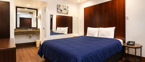 Habitación de hotel con una cama grande, cabecera de madera, mesita de noche con teléfono, y baño con espejo y lavamanos.