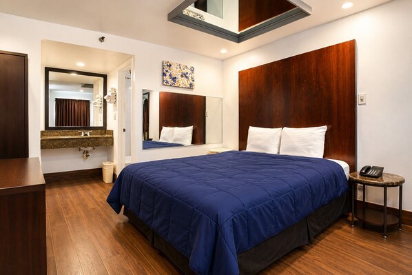 A hotel room with a large bed, a wooden headboard, a bedside table with a phone, and a bathroom with a mirror and sink.