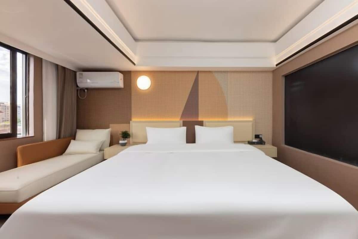 A hotel room with a large bed, a sofa, a bedside table, an air conditioning unit, and a large window.