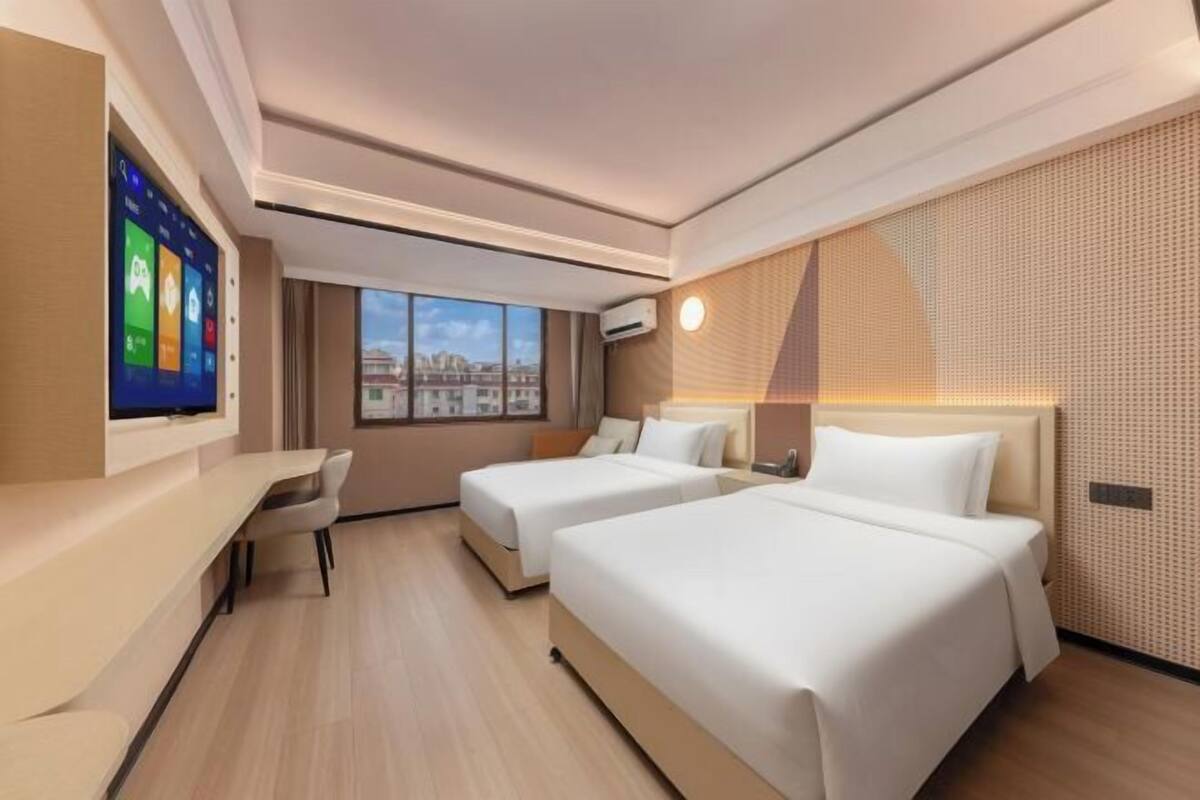 A hotel room with two beds, a desk, a TV, and a window with a city view.
