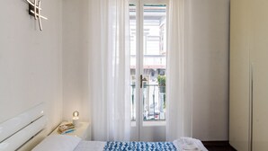 A hotel room with a bed, a nightstand with books, a window with curtains, and a view of the street outside. - Casa Fubis (Viareggio)