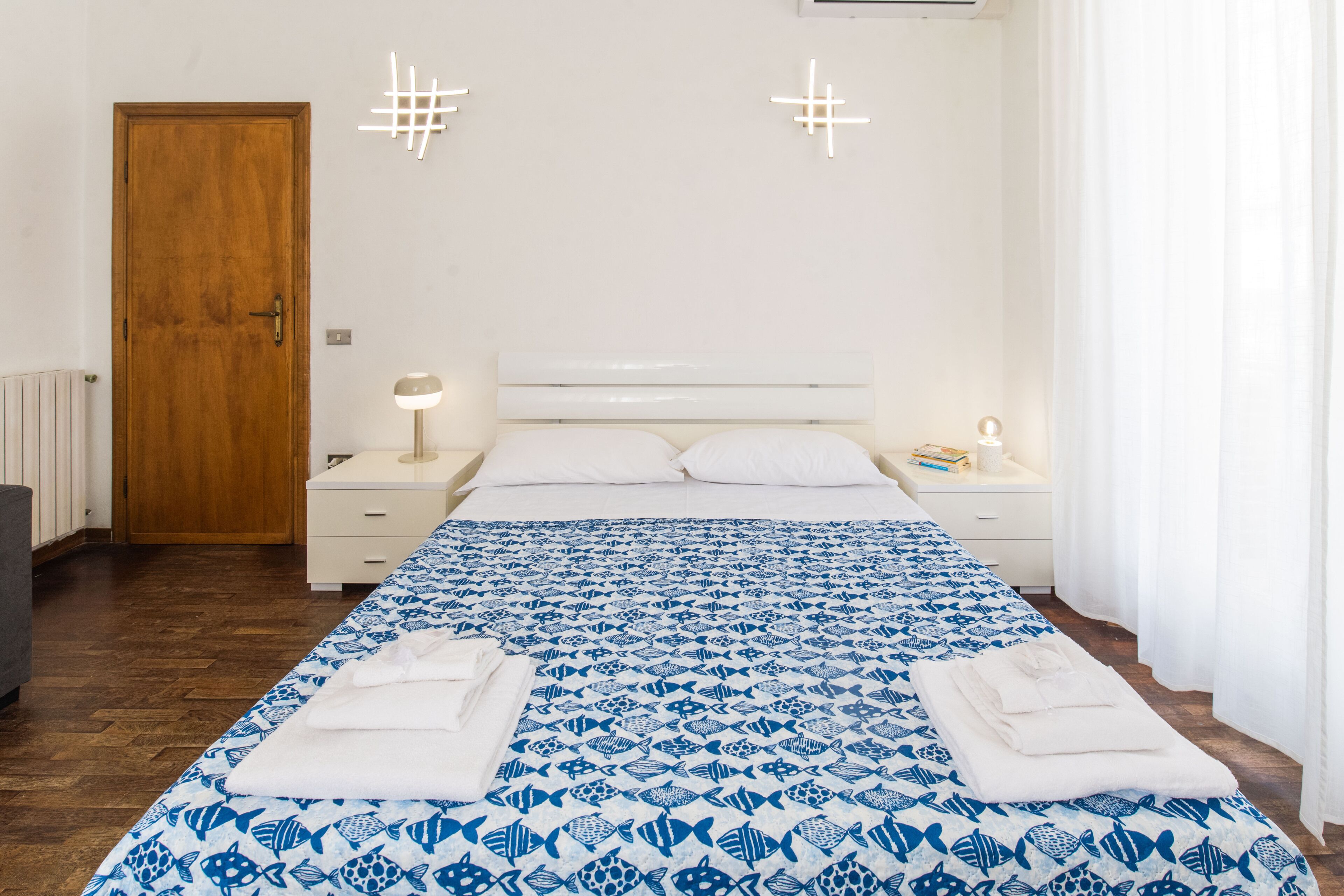 A bedroom with a wooden door, white bedside tables, a bed with a blue and white patterned bedspread, and white curtains.