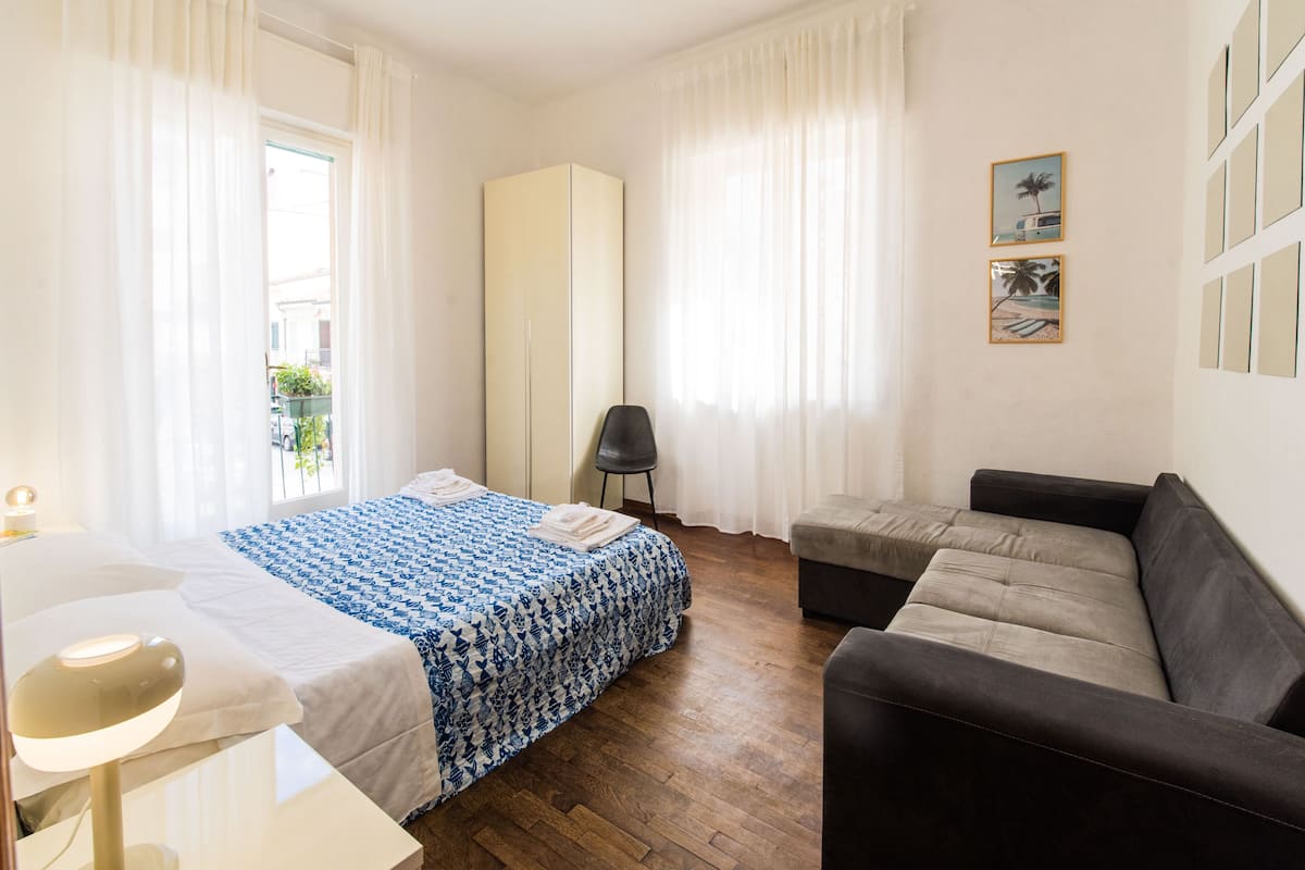 A hotel room with a bed, a sofa, a desk with a lamp, and a window with curtains.