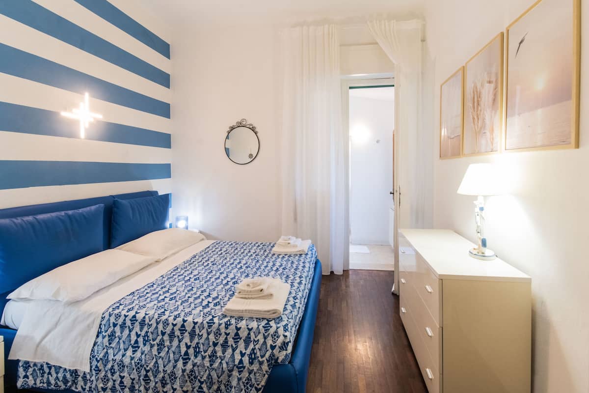 A hotel room with a bed, a bedside table with a lamp, a mirror, and a door leading to another room.