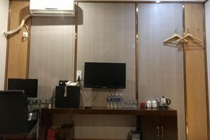 Habitación de hotel con televisor de pantalla plana en la pared, un escritorio de madera con computadora y parlantes, un mueble con dispensador de agua y un armario con perchas de madera.