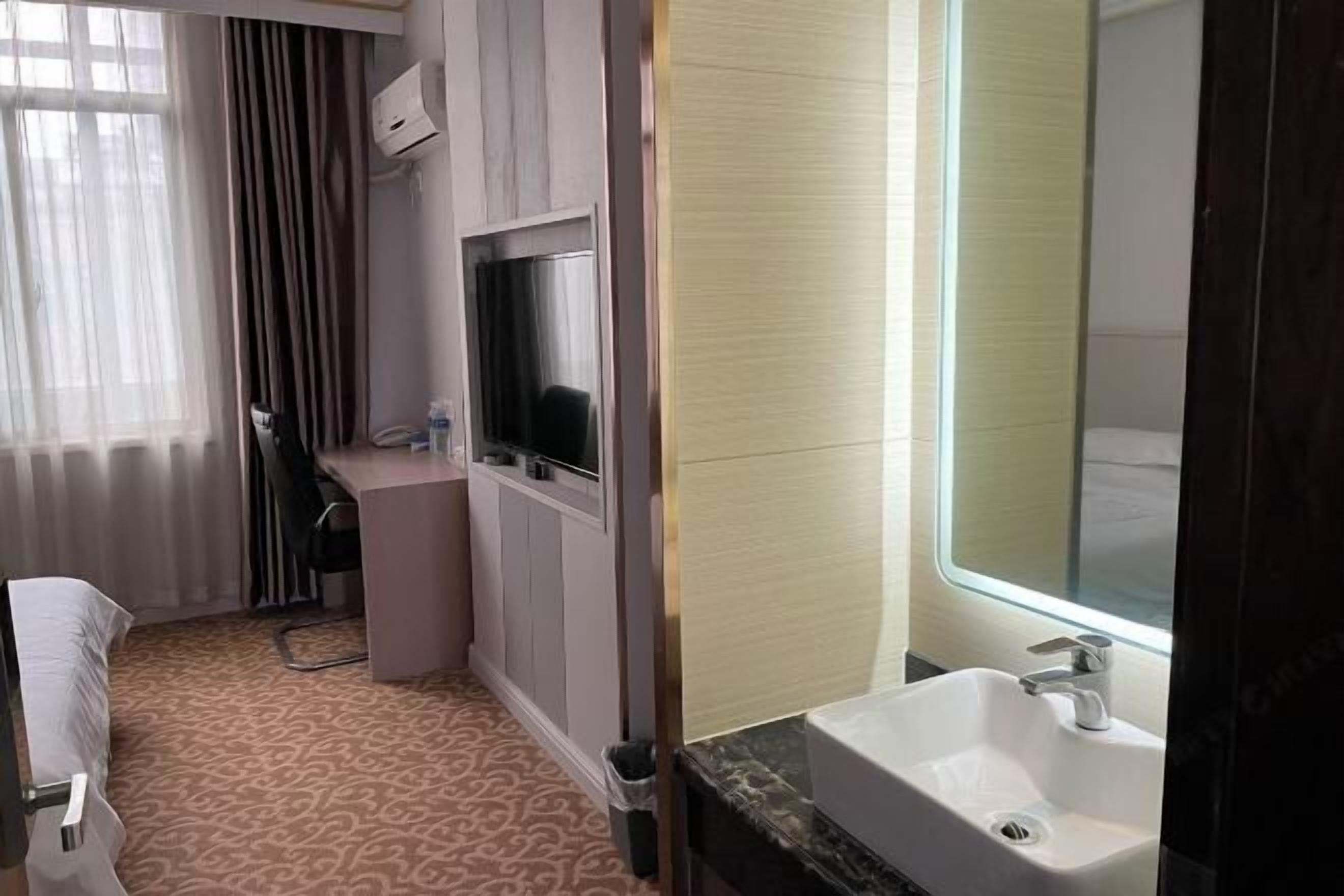 A hotel room with a bed, a desk with a chair, a TV, a bathroom with a sink and mirror, and a patterned carpet.