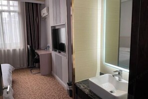 Habitación de hotel con cama, escritorio con silla, televisor, baño con lavamanos y espejo, y alfombra estampada.