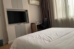 Habitación de hotel con una cama grande, un televisor de pantalla plana montado en la pared, un escritorio con una silla y una ventana con cortinas translúcidas.