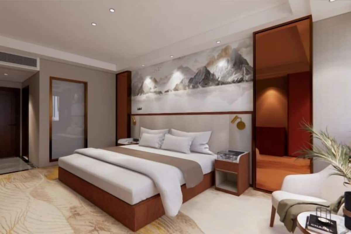 A modern hotel room with a large bed, a nightstand, a seating area, and a wall with a mountain landscape painting.