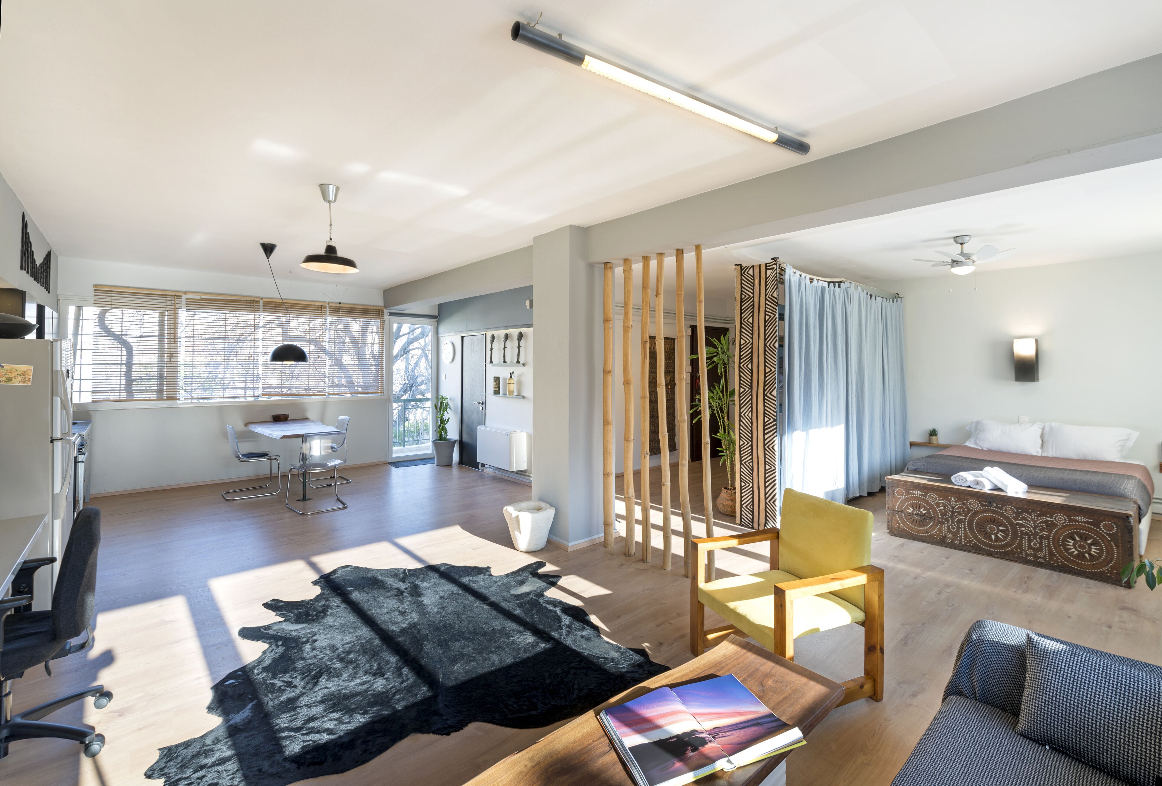 Uma sala de estar moderna com sofá, mesa de centro e área de jantar com mesa e cadeiras. Uma cama com cabeceira estampada aparece ao fundo.