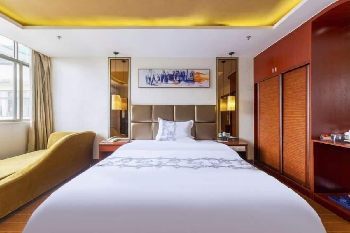A hotel room with a large bed, a bedside table, a nightstand, a chair, a wardrobe, and a painting on the wall.