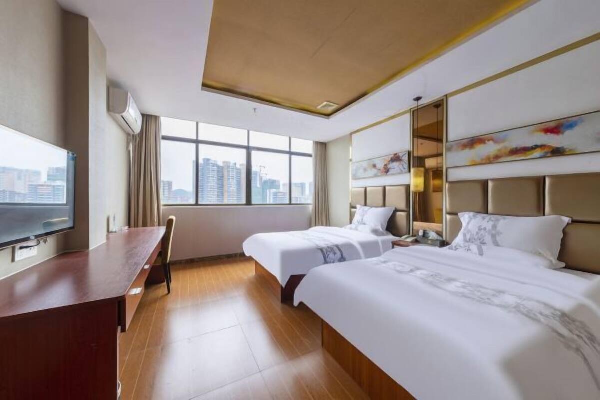 A hotel room with two beds, a desk, a television, and a large window with a city view.