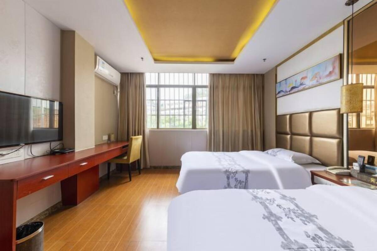 A hotel room with a wooden desk, two beds, a TV, and a window with curtains.