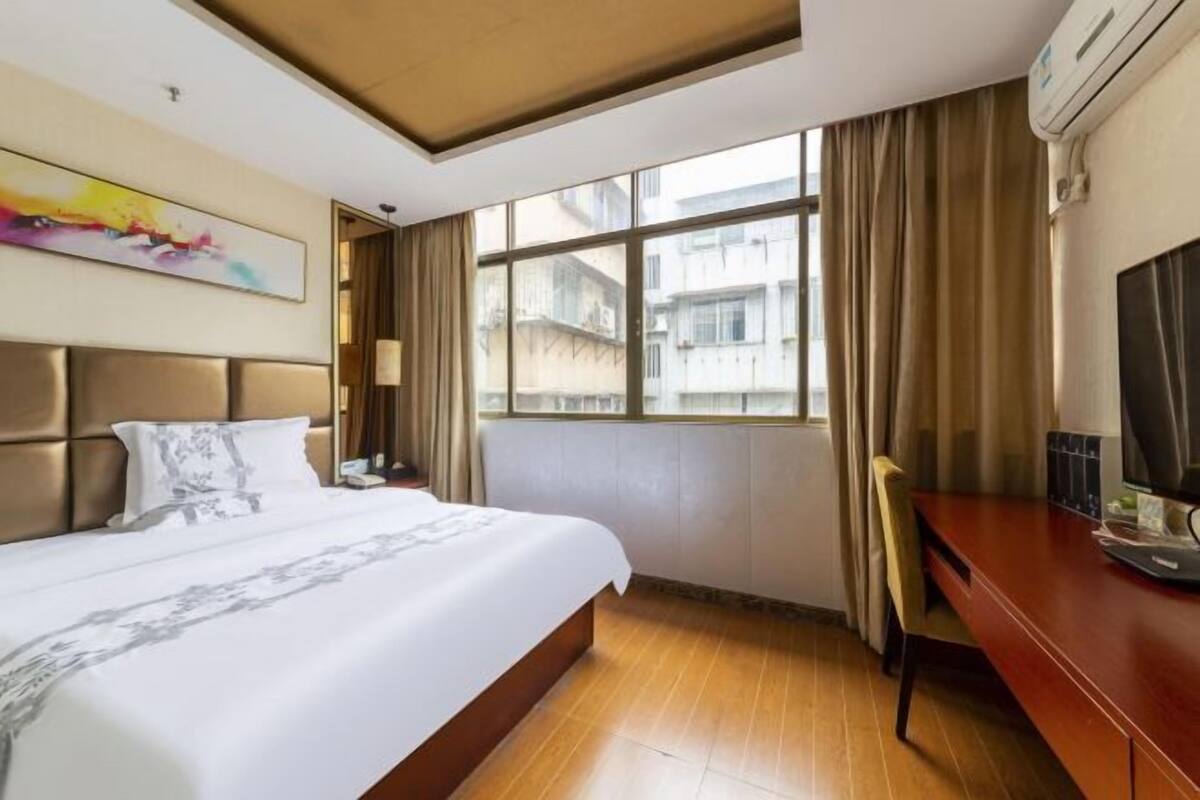 A hotel room with a bed, a desk, a chair, a window with curtains, a painting on the wall, and an air conditioning unit.