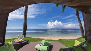 Vue sur la plage depuis une tente, avec un espace herbeux, une table en osier sur laquelle se trouve un panier de fruits et des verres, et une serviette verte.