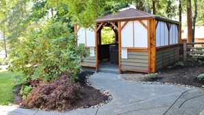 A wooden structure with a sloped roof and a door, surrounded by a landscaped garden with a small tree and mulch.