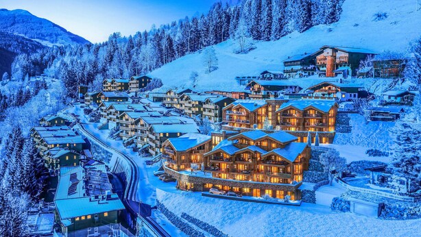 Un complejo turístico en la montaña cubierto de nieve, con varios edificios de madera, incluyendo un destacable hotel con balcones y un telesilla visible al fondo.