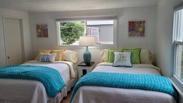 A bedroom with two beds, each with a blue blanket, white bedding, and decorative pillows. There is a window with a view of greenery outside.