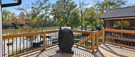 A wooden deck with a grill, overlooking a lake with trees and a dock.