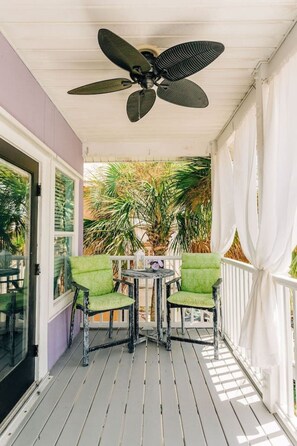 Un espace extérieur abrité, doté d’un ventilateur de plafond, de chaises vertes, d’une petite table et offrant une vue sur des palmiers.