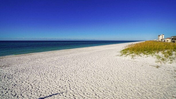 Une plage de sable blanc, avec une eau d’un bleu limpide et des bâtiments visibles à l’arrière-plan.