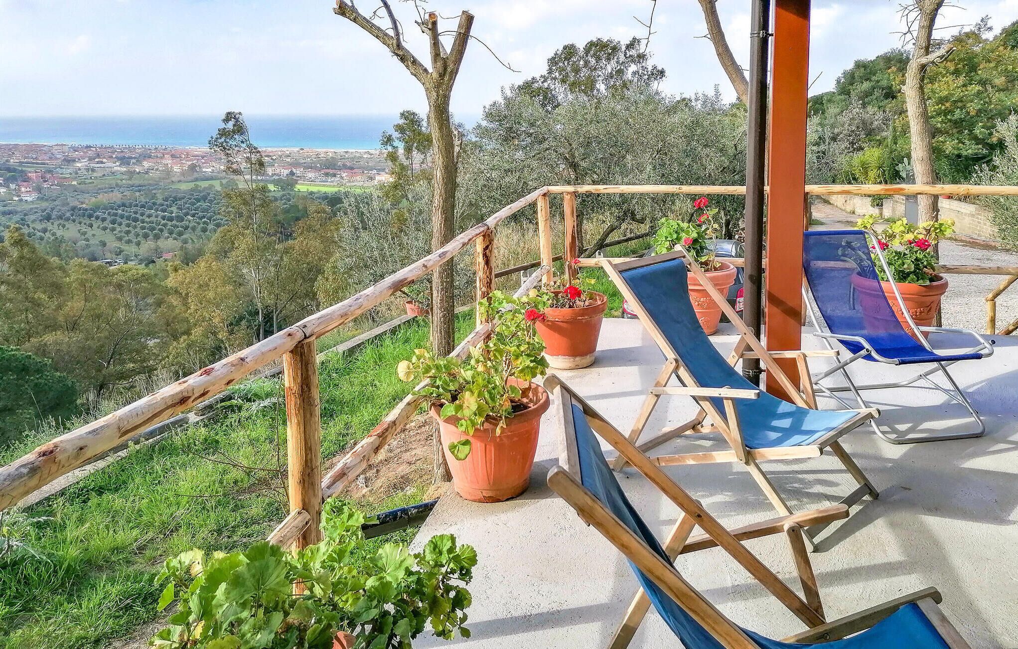 Terraço com guarda-corpos de madeira, plantas em vasos e espreguiçadeiras azuis com vista para a paisagem costeira.
