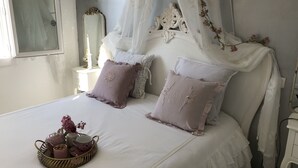 A bedroom with a white bed, decorative pillows, a bedside table with a lamp, and a window with sheer curtains. - Small warm house. (Le Château-d'Oléron)