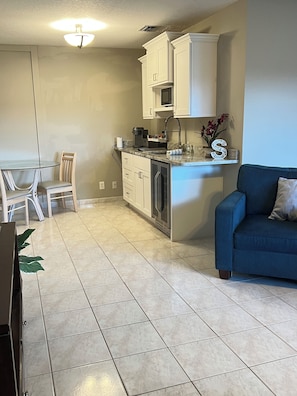 A compact kitchen with white cabinets, a microwave, and a coffee maker. There is a dining area with a glass table and chairs, and a blue sofa in the living area. - Wellington 1bed 1bath, Just minutes to WEF and Polo grounds (Wellington)