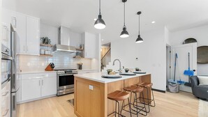 Uma cozinha moderna com armários brancos, forno de aço inoxidável, ilha de madeira com banquetas e iluminação pendente.