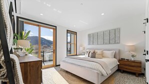 Quarto com uma cama grande, uma mesa de cabeceira de madeira, uma luminária no teto e uma janela ampla com vista para montanhas.
