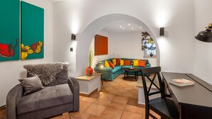 A modern living room with a grey sofa, a teal sofa with colorful cushions, a dining table with chairs, and a large window with a view of the city.