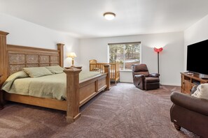 Une chambre à coucher avec un lit en bois, un fauteuil inclinable, un téléviseur et une fenêtre donnant sur des arbres.