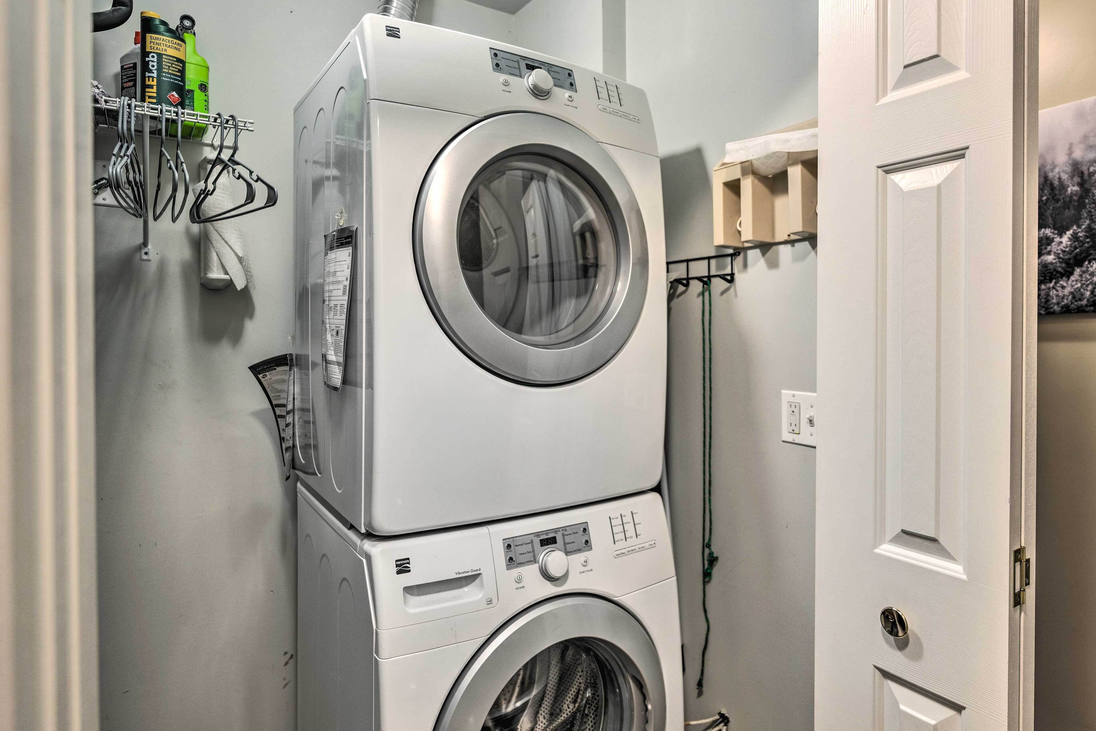 Un appareil combiné lave-linge et sèche-linge, installé en hauteur, se trouve dans une buanderie. Une porte est située à droite, et un porte-serviettes est placé au-dessus du sèche-linge.