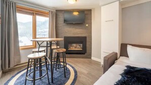 Moderni hotellihuone, jossa on takka, seinälle asennettu televisio, pehmeällä heitteellä varustettu sänky ja suuret ikkunat, joista avautuu näkymä ulos.