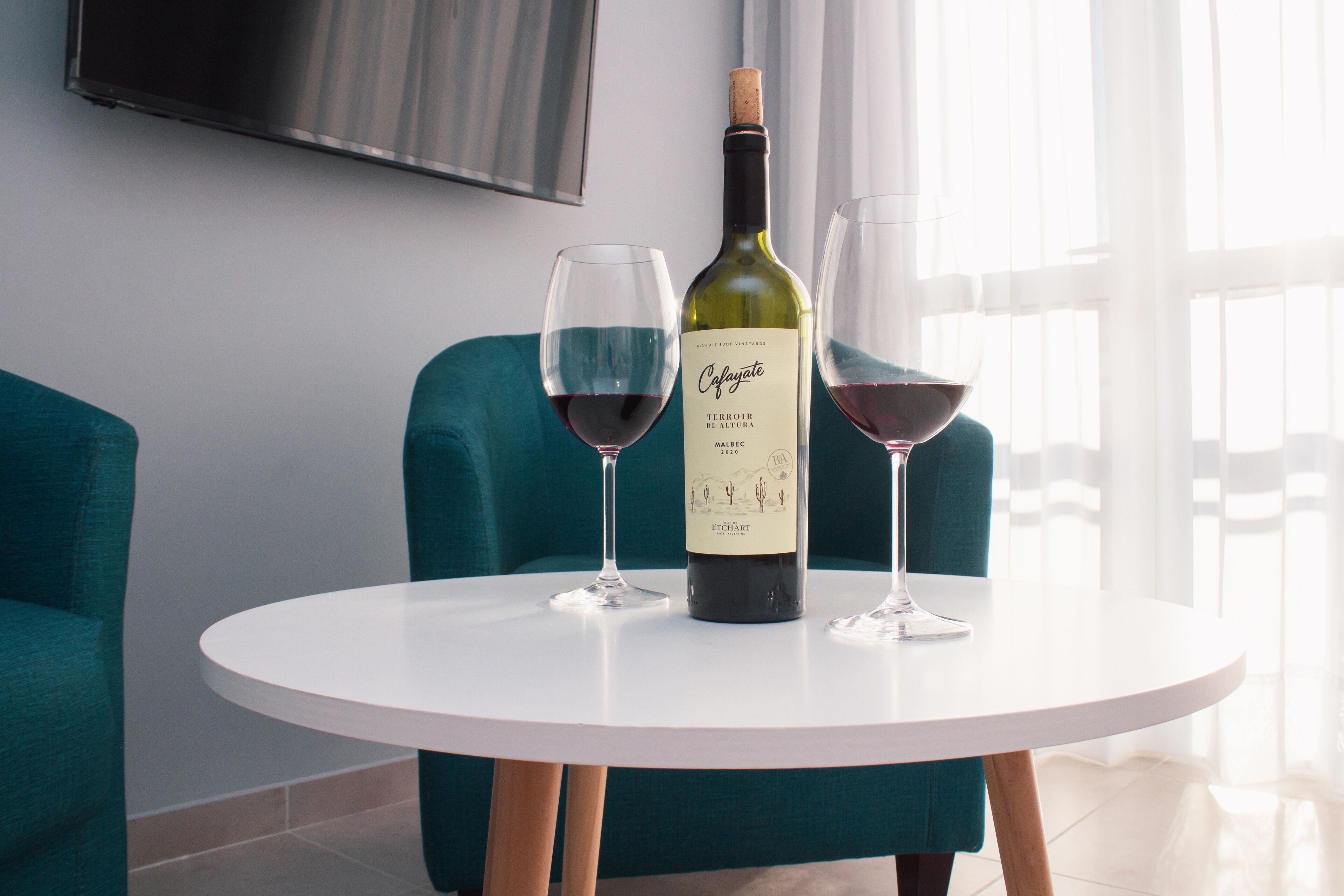 A round white table with a bottle of wine and two glasses on it, set against teal chairs and a white wall with a mounted TV.