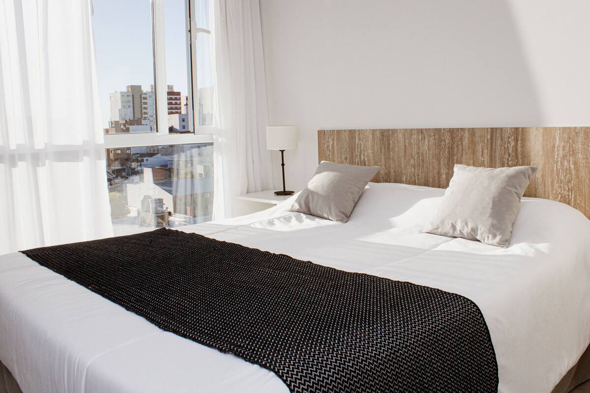 Une chambre d’hôtel avec un grand lit, du linge de lit blanc, une vue sur la ville par la fenêtre et une tête de lit en bois.
