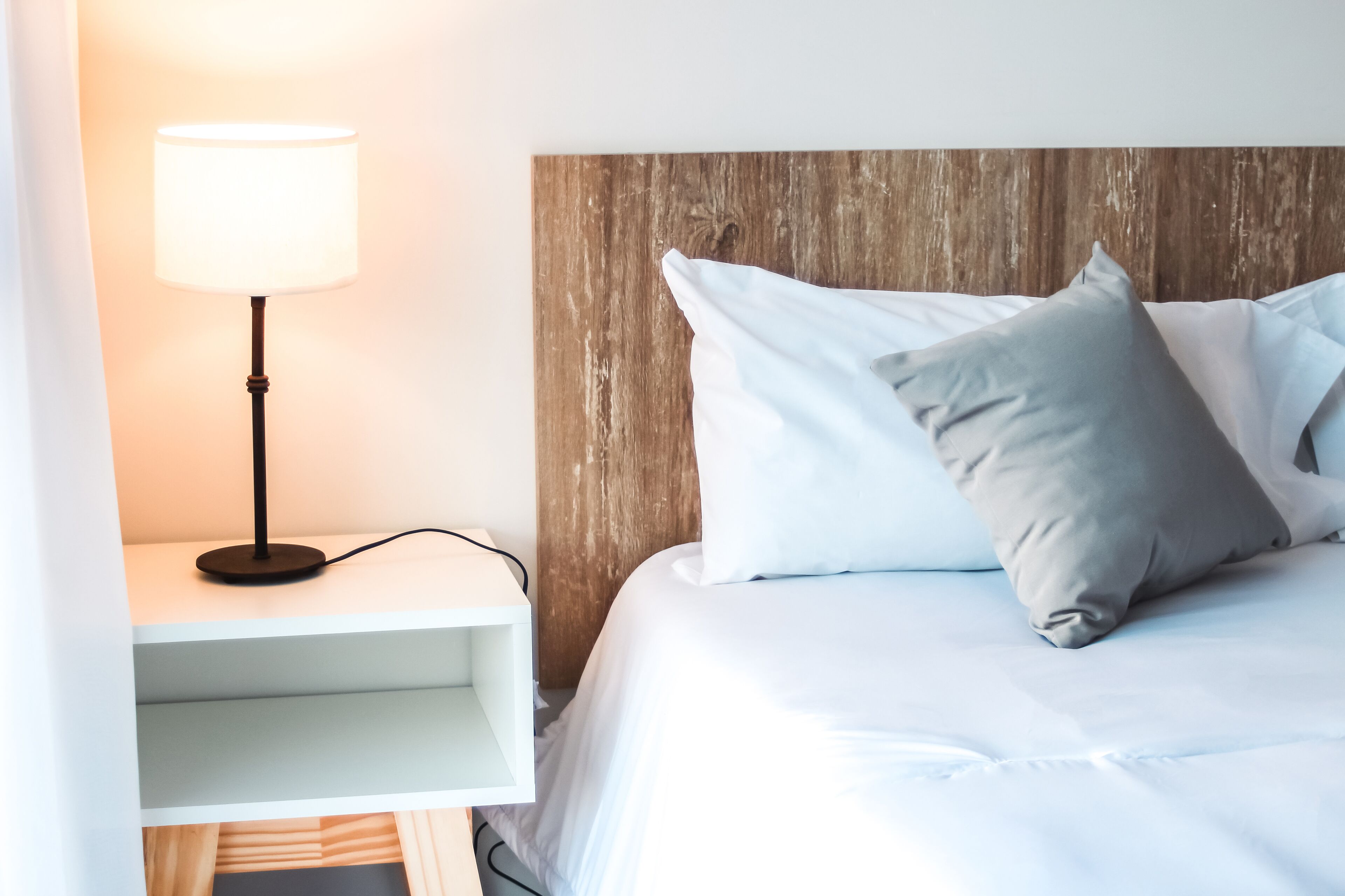 A neatly made bed with white and grey pillows, a wooden headboard, and a bedside table with a lamp.