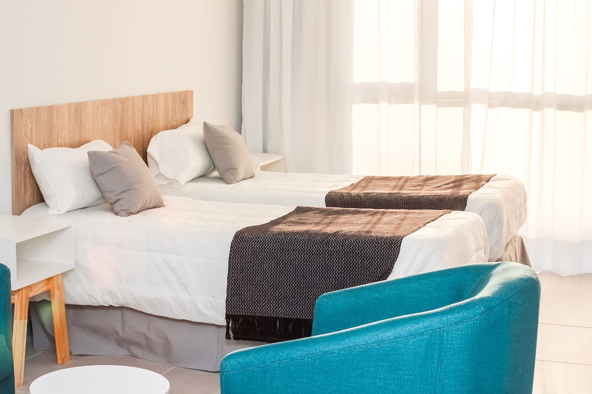 Une chambre d’hôtel avec deux lits, une tête de lit en bois, un bureau blanc et des chaises de couleur turquoise.