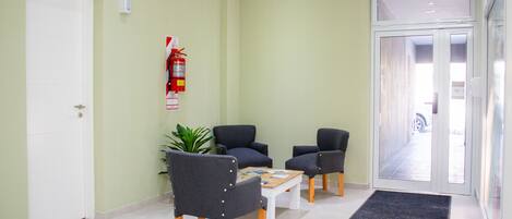 Un área de recepción con una puerta blanca, un extintor en la pared, dos sillones, una mesita y un felpudo en la entrada.