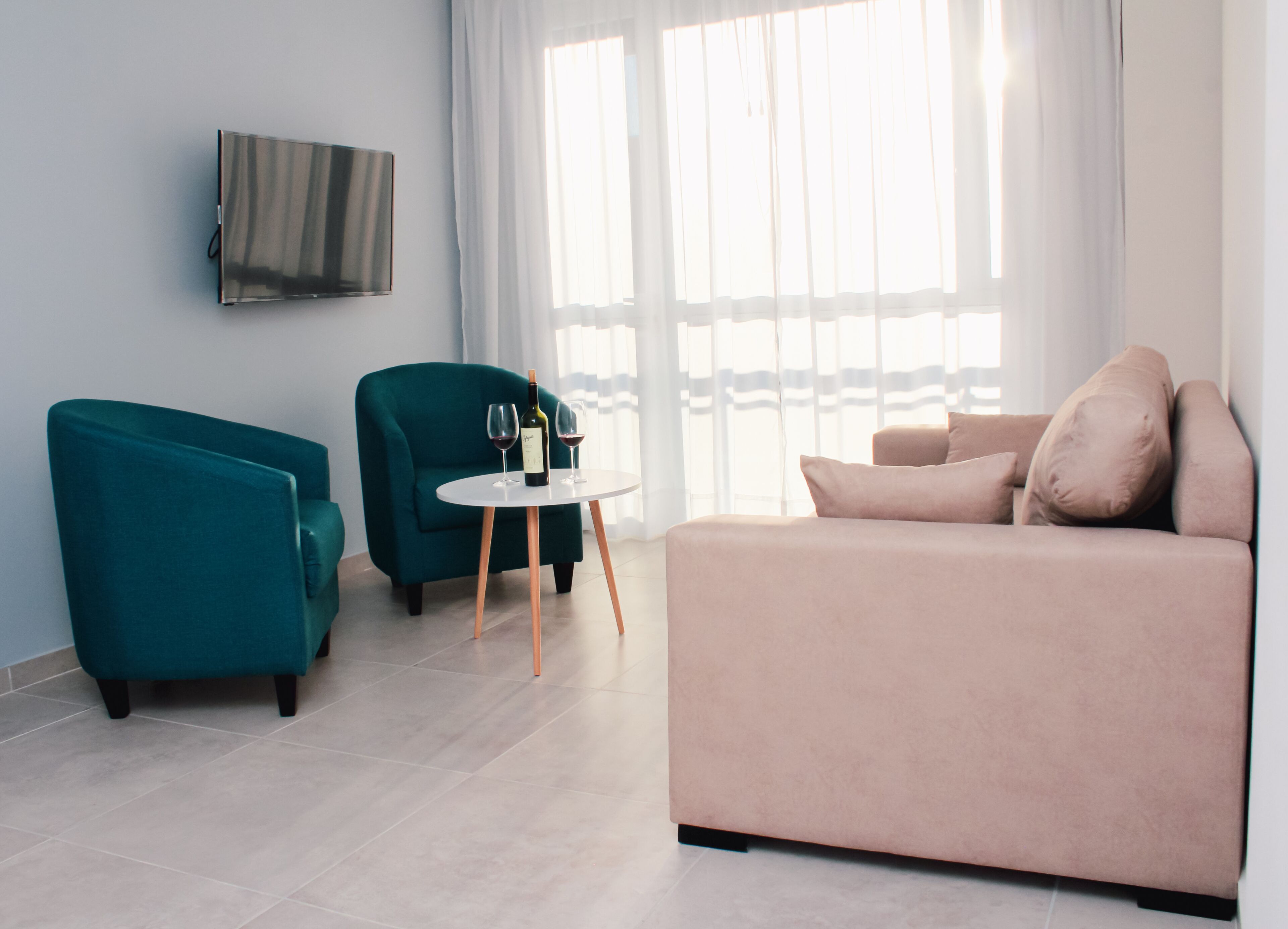Uma sala de estar moderna com sofás verde-água e bege, uma mesa redonda pequena com copos de vinho, e uma TV de tela plana na parede.
