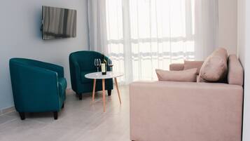 Uma sala de estar moderna com sofás verde-água e bege, uma mesa redonda pequena com copos de vinho, e uma TV de tela plana na parede.