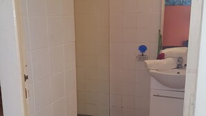 A bathroom with white tiled walls, a shower with a glass door, a sink with a mirror, and a small orange rug. - Emerald b (Sainte-Rose)